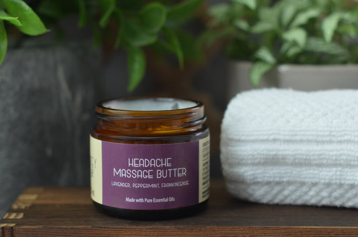 Open jar of Kinsley Oils Headache Massage Butter with lavender, peppermint, and frankincense essential oils, displayed on a wooden shelf beside a folded white towel and soft greenery.