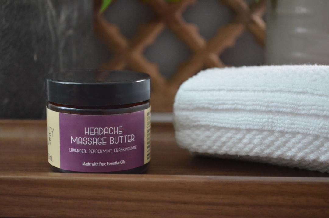 Kinsley Oils Headache Massage Butter with lavender, peppermint, and frankincense essential oils in an amber jar, displayed on a wooden surface beside a folded white towel.