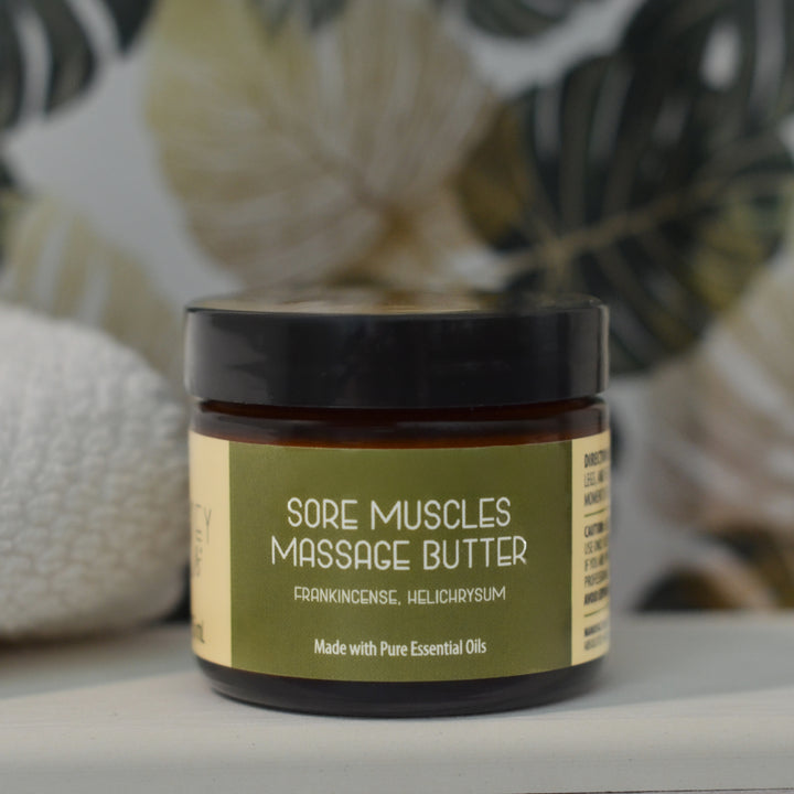 Kinsley Oils Sore Muscles Massage Butter with frankincense and helichrysum essential oils in an amber jar with a black lid, displayed beside a rolled white towel against a softly blurred botanical background.
