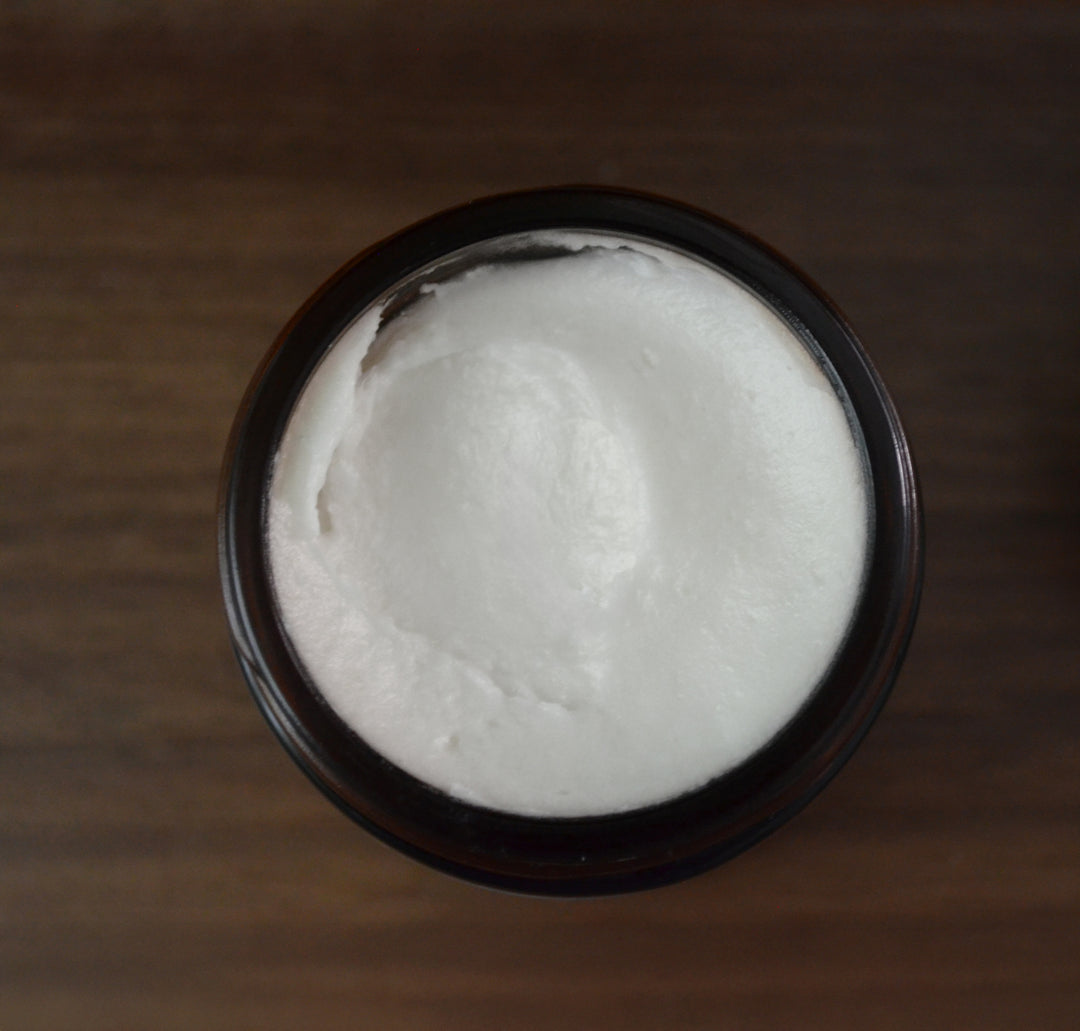 Overhead view of an open amber jar filled with rich, creamy white Kinsley Oils Massage Butter, showcasing its smooth, buttery texture against a dark wooden background.