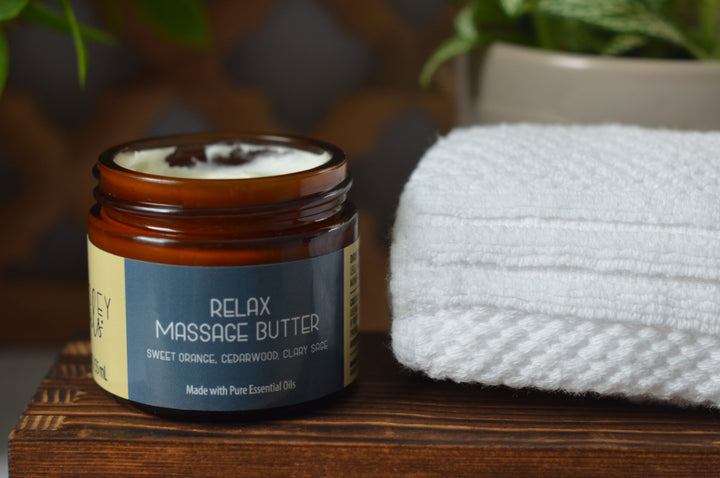 Open amber jar of Kinsley Oils Relax Massage Butter with sweet orange, cedarwood, and clary sage essential oils, displayed on a wooden shelf beside a folded white towel and softly blurred greenery.