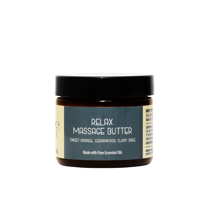 Front view of Kinsley Oils Relax Massage Butter with sweet orange, cedarwood, and clary sage essential oils, packaged in an amber jar with a black lid and blue-gray label against a white background.