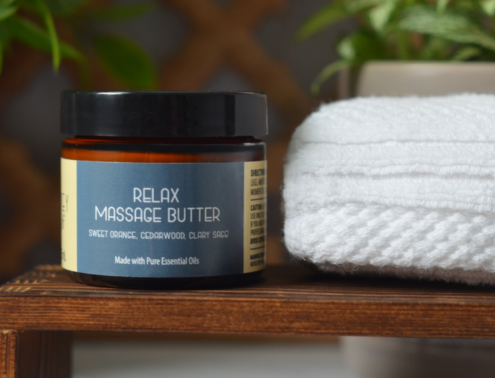 Kinsley Oils Relax Massage Butter with sweet orange, cedarwood, and clary sage essential oils in an amber jar with a black lid, displayed on a wooden shelf beside a folded white towel and greenery.