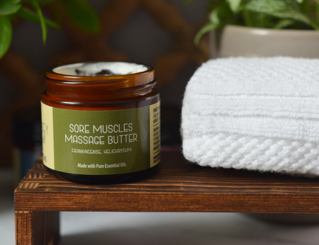 Open amber jar of Kinsley Oils Sore Muscles Massage Butter with frankincense and helichrysum essential oils, displayed on a wooden shelf beside a folded white towel and softly blurred greenery.