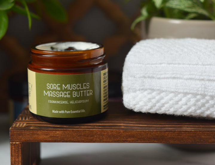 Open amber jar of Kinsley Oils Sore Muscles Massage Butter with frankincense and helichrysum essential oils, displayed on a wooden shelf beside a folded white towel and softly blurred greenery.